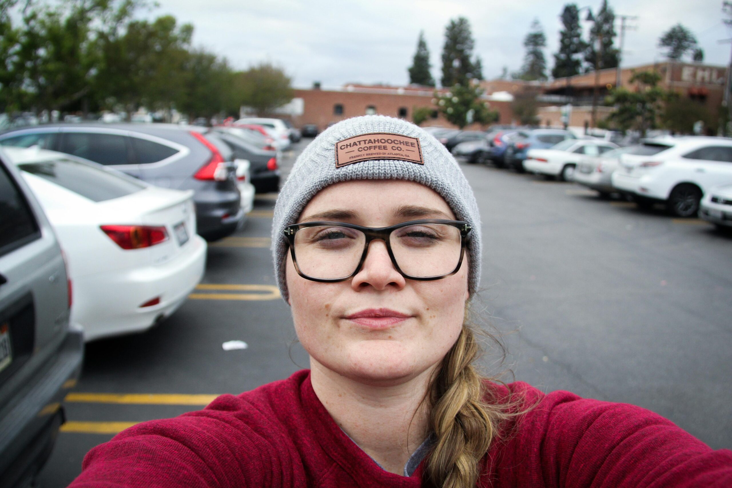 Portrait of a woman wearing glasses and a beanie, taking a selfie in a busy parking lot.