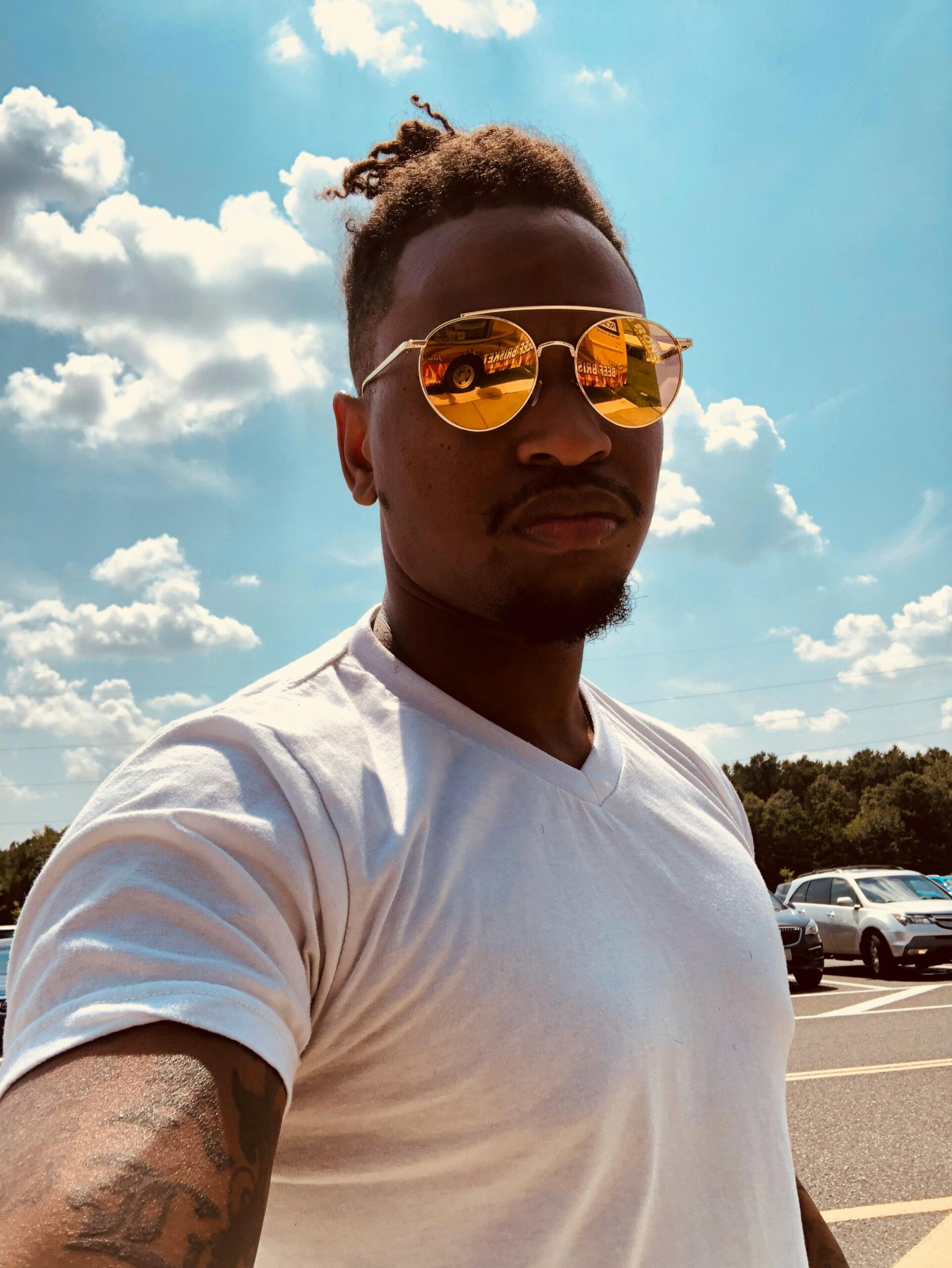 Young man in reflective sunglasses taking a selfie on a sunny day with clear skies and parked cars in the background.